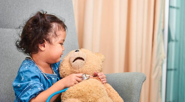 Cute curly haired Asian girl, about 3 years old with stethoscope plays with teddy bear while sitting on the chair in the living room. - Foto, imagen