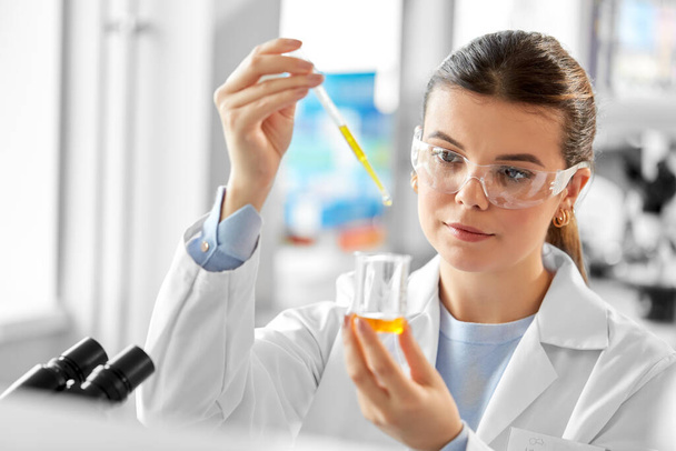 science research, work and people concept - female scientist in goggles with dropper and chemical or test sample in beaker working in laboratory - Φωτογραφία, εικόνα