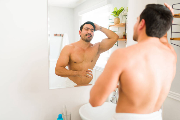 Attractive hispanic young man looking in the bathroom mirror after a shower and getting ready while putting hair gel  - Foto, imagen