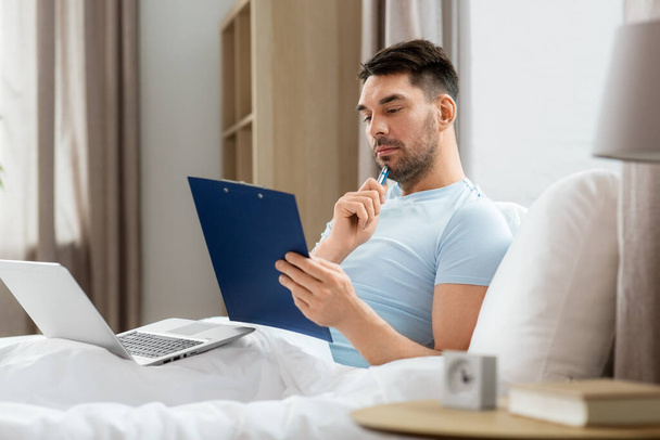 people, technology and remote job concept - man with laptop computer and clipboard working in bed at home bedroom - Photo, Image