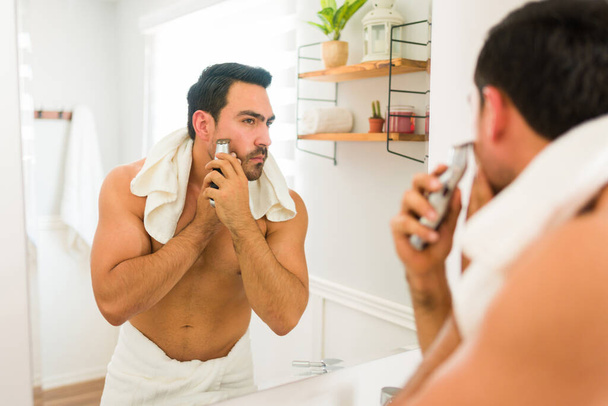 М'язистий молодий чоловік використовує рушник і бороду, готуючись у дзеркалі ванної кімнати
 - Фото, зображення