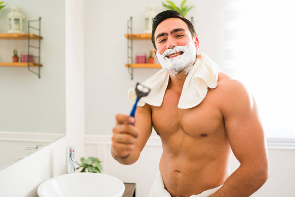 Cheerful muscular man with shaving cream on his face using a disposable razor to shave his facial hair before showering - Zdjęcie, obraz