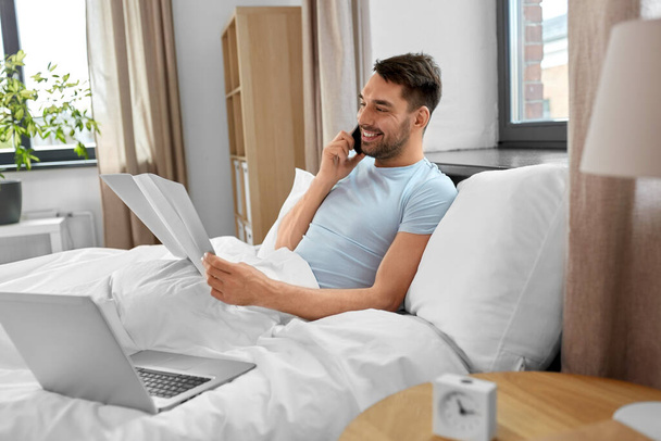 people, technology and remote job concept - man with folder and laptop computer calling on smartphone in bed at home bedroom - Фото, изображение