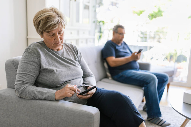 Senior couple sitting on couch and using their mobile phones at home - Photo, Image