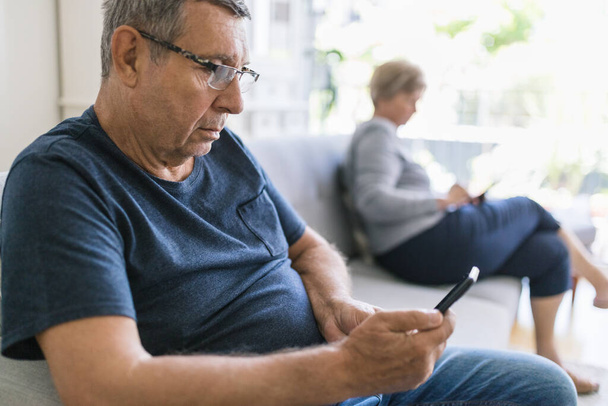 Senior couple sitting on couch and using their mobile phones at home - Foto, Imagem