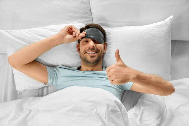 people, bedtime and rest concept - happy smiling man in sleeping eye mask lying in bed and showing thumbs up gesture, top view - Photo, image