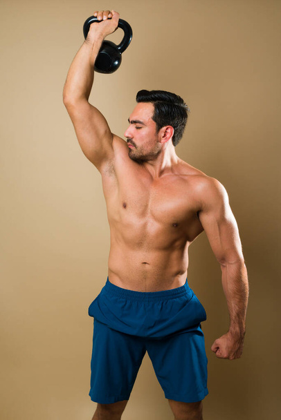 Сильный кавказский молодой человек поднимает гирь и смотрит на свои бицепсные мышцы во время тренировки  - Фото, изображение