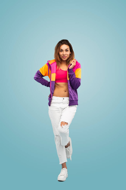 Full length of confident young female millennial with long brown hair in trendy crop top and jacket, standing against blue background with hand on waist and looking at camera - Foto, immagini