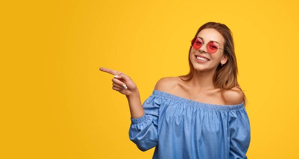 Delighted young female model with long dark hair in blouse and sunglasses smiling at camera and pointing aside against yellow background - Foto, immagini