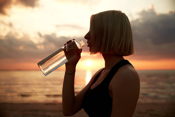 fitness, sport, and healthy lifestyle concept - woman drinking water from bottle on beach over sunset - 写真・画像