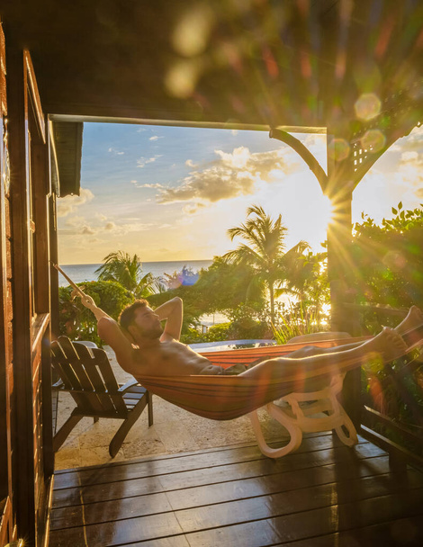 Young men in swim shorts sunbathing in a hammock at Saint Lucia Caribbean, men at infinity pool during sunset. man in swim shorts at the pool - Photo, Image