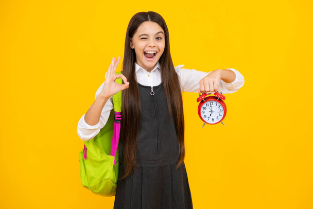 Funny face. Back to school. Teenager school girl with backpack hold clock alarm, time to learn. School children on isolated yellow background - Photo, Image