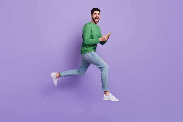 Full body photo of young latin guy hold device impressed repost like jumping wear stylish green clothes isolated on purple color background. - Valokuva, kuva