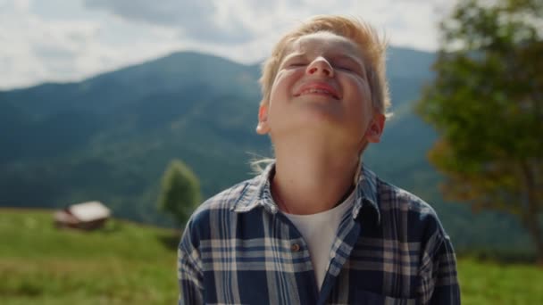 Closeup little boy enjoying sunlight raising head to sky in summer mountains. Cute child relaxing on green meadow sunny day. Portrait of happy smiling guy standing alone outdoors on beautiful nature.  - Filmati, video