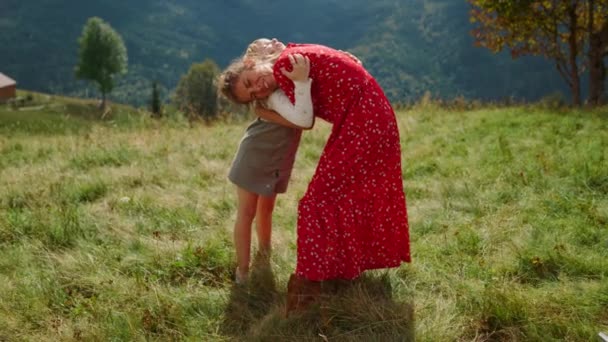 Happy woman with daughter hugging on green meadow sunny day. Smiling mother with lovely child enjoying summer holiday in mountains. Cheerful mom with cute girl spending time together outdoors. - Materiał filmowy, wideo