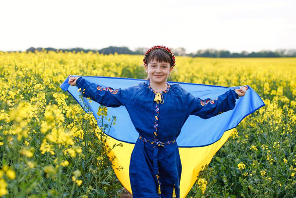 Betet für die Ukraine. Kind mit ukrainischer Flagge im Rapsfeld. Ein Mädchen im bestickten Hemd läuft über das Feld, die ukrainische Flagge in der Hand - Foto, Bild