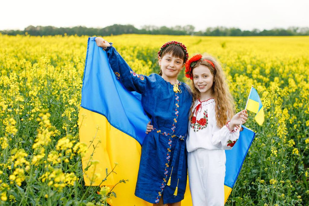 Pray for Ukraine. Two cute girls holding in hands a blue and yellow Ukrainian national flag in the middle of a rapeseed field - Foto, imagen