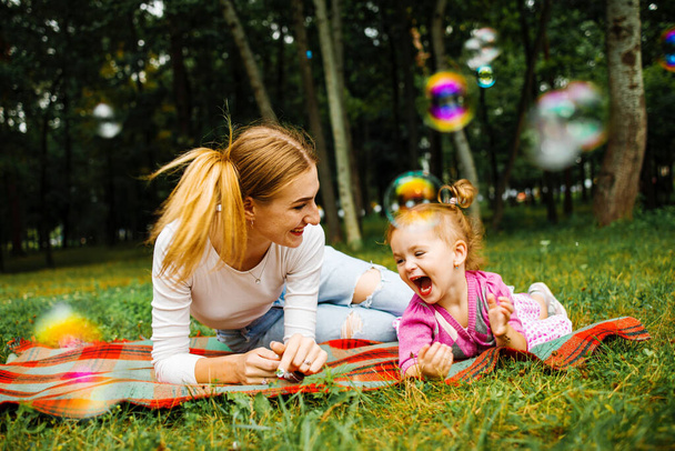 Молода мати зі своєю милою дівчинкою дме мильні бульбашки в літньому парку
. - Фото, зображення