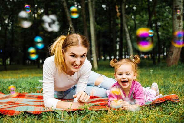 Young mother with her cute little girl is blowing a soap bubbles in summer park. - Fotó, kép