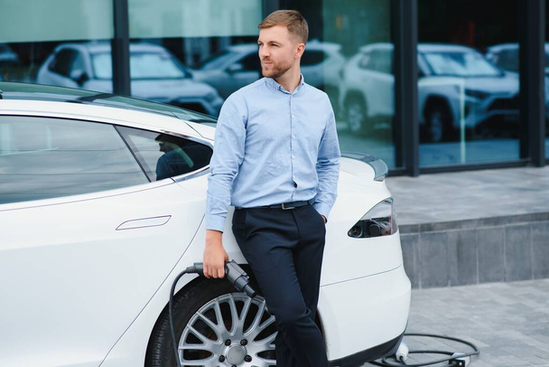 portrait of young handsome man in casual wear, standing at the charging station. Eco electric car concept - Φωτογραφία, εικόνα