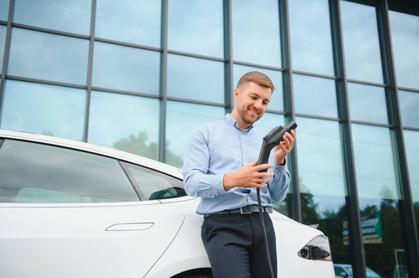 Casual man with smartphone near electric car waiting for the finish of the battery charging process. - Foto, Bild