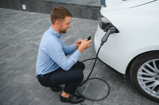 Man charging his electric car at charge station and using smartphone. - Zdjęcie, obraz