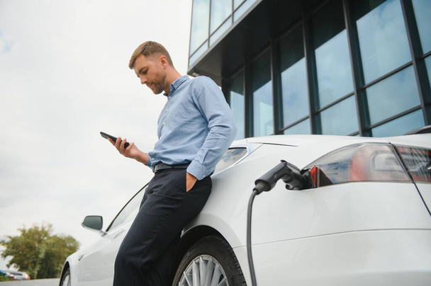 Man charging his electric car at charge station and using smartphone. - Valokuva, kuva