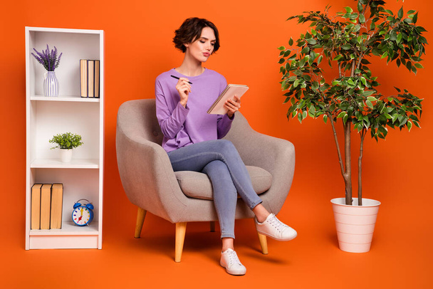 Full size portrait of focused intelligent lady sitting cozy chair hold notepad isolated on orange color background. - Фото, изображение
