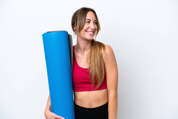 Young sport woman going to yoga classes while holding a mat isolated on white background looking to the side and smiling - Fotoğraf, Görsel