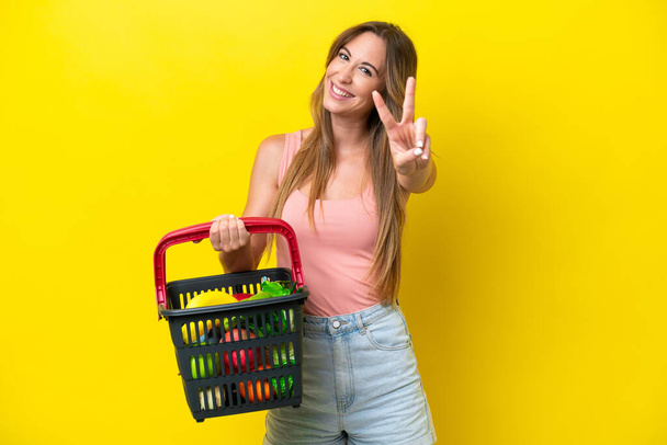 黄色の背景の笑顔で孤立した食べ物でいっぱいのショッピングバスケットを保持し、勝利の兆候を示す若い白人女性 - 写真・画像