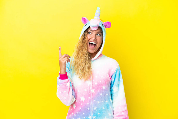 Girl with curly hair wearing a unicorn pajama isolated on yellow background pointing up and surprised - Foto, immagini