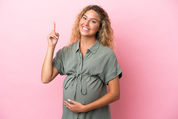 Meisje met krullend haar geïsoleerd op roze achtergrond zwanger en wijzen omhoog - Foto, afbeelding