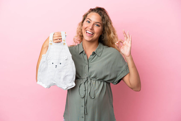 Girl with curly hair isolated on pink background pregnant and holding baby clothes with ok sign - Foto, Bild