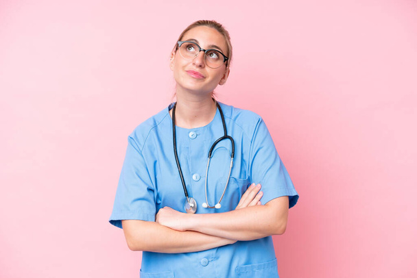 Joven enfermera cirujana aislada sobre fondo rosa mirando hacia arriba mientras sonríe - Foto, Imagen