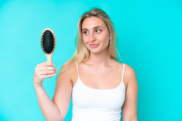 Young caucasian woman with hair comb isolated on blue background with happy expression - Foto, afbeelding
