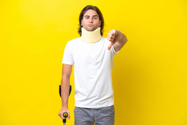 Young handsome man wearing neck brace and crutches isolated on yellow background showing thumb down with negative expression - Valokuva, kuva