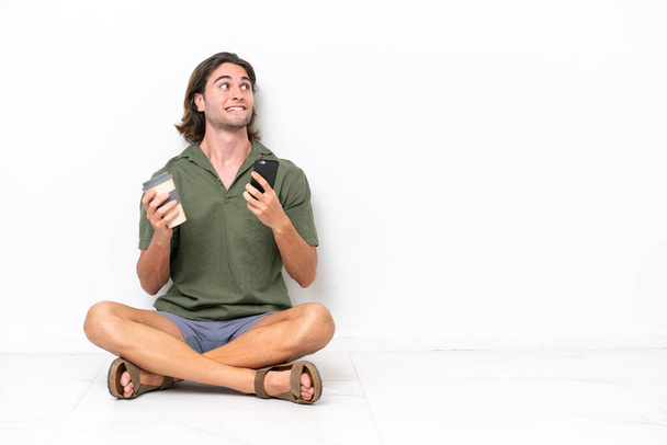 Young handsome man sitting on the floor isolated on white background holding coffee to take away and a mobile while thinking something - Photo, image