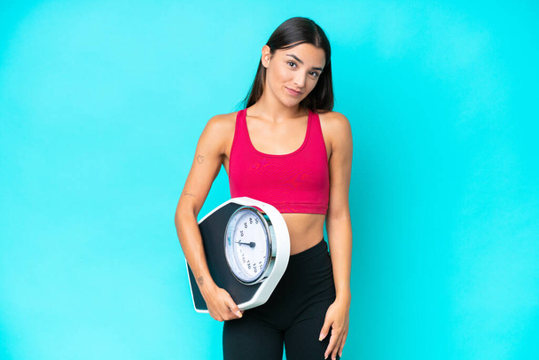 Young caucasian woman isolated on blue background with weighing machine - Foto, Imagen