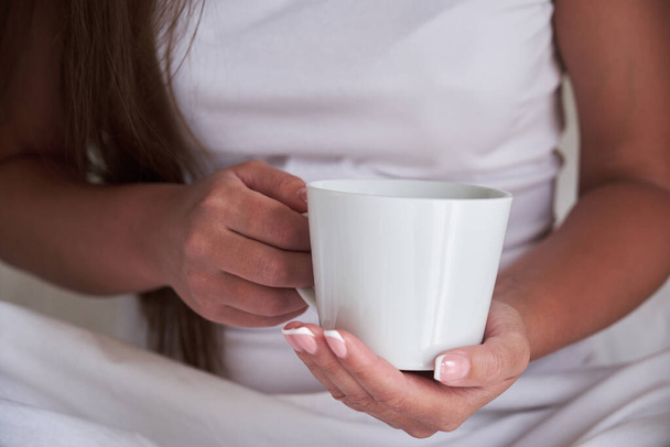 Close-up of female hands with a cup of drink. A beautiful girl is holding a cup of tea or coffee while sitting in bed in the morning. Mockup for your design. - Фото, зображення
