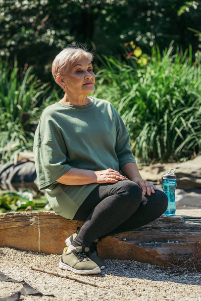 retired woman in sportswear sitting with sports bottle on wooden log around green plants - Foto, imagen