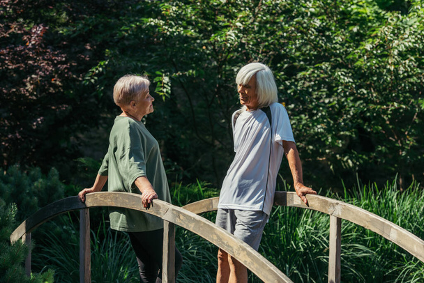 retired senior couple in sportswear looking at each other while standing on bridge  - Foto, immagini