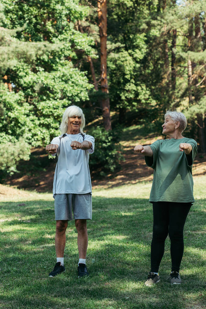 full length of joyful senior couple in sportswear exercising with outstretched hands together in park - Photo, image