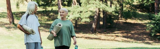 cheerful senior couple in sportswear holding fitness mats and sports bottles in park, banner - Foto, imagen