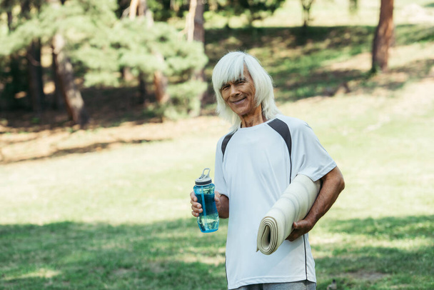 happy senior man in white t-shirt holding sports bottle and fitness mat in park - Foto, afbeelding