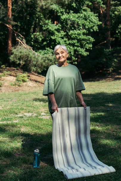 full length of happy senior woman with grey hair holding fitness mat in park  - Photo, Image