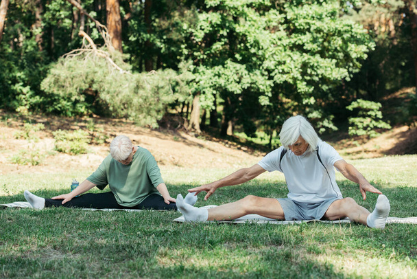 full length of senior couple stretching while exercising on fitness mats in green park - Foto, Imagen