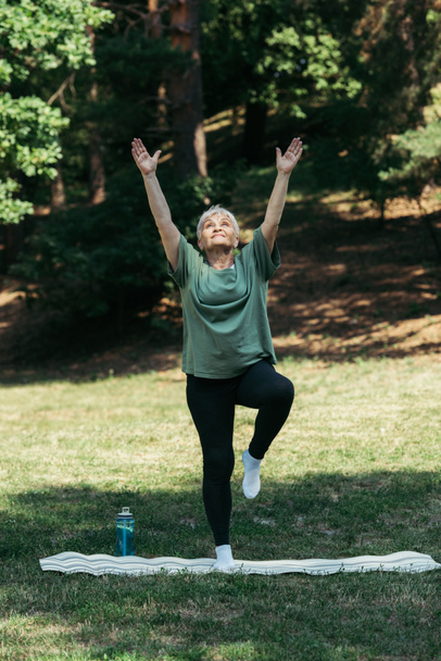 full length of senior woman with raised hands exercising on fitness mat in park - Foto, afbeelding