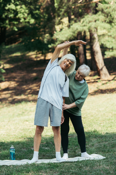 senior woman training happy husband with grey hair standing on fitness mat in park - Foto, afbeelding