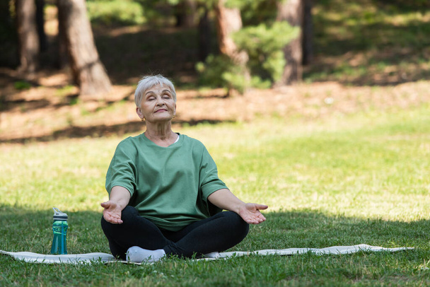 full length of senior woman with grey hair meditating on fitness mat in park - Zdjęcie, obraz