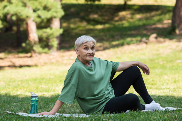 full length of senior woman sitting on fitness mat near sports bottle in park - Foto, imagen